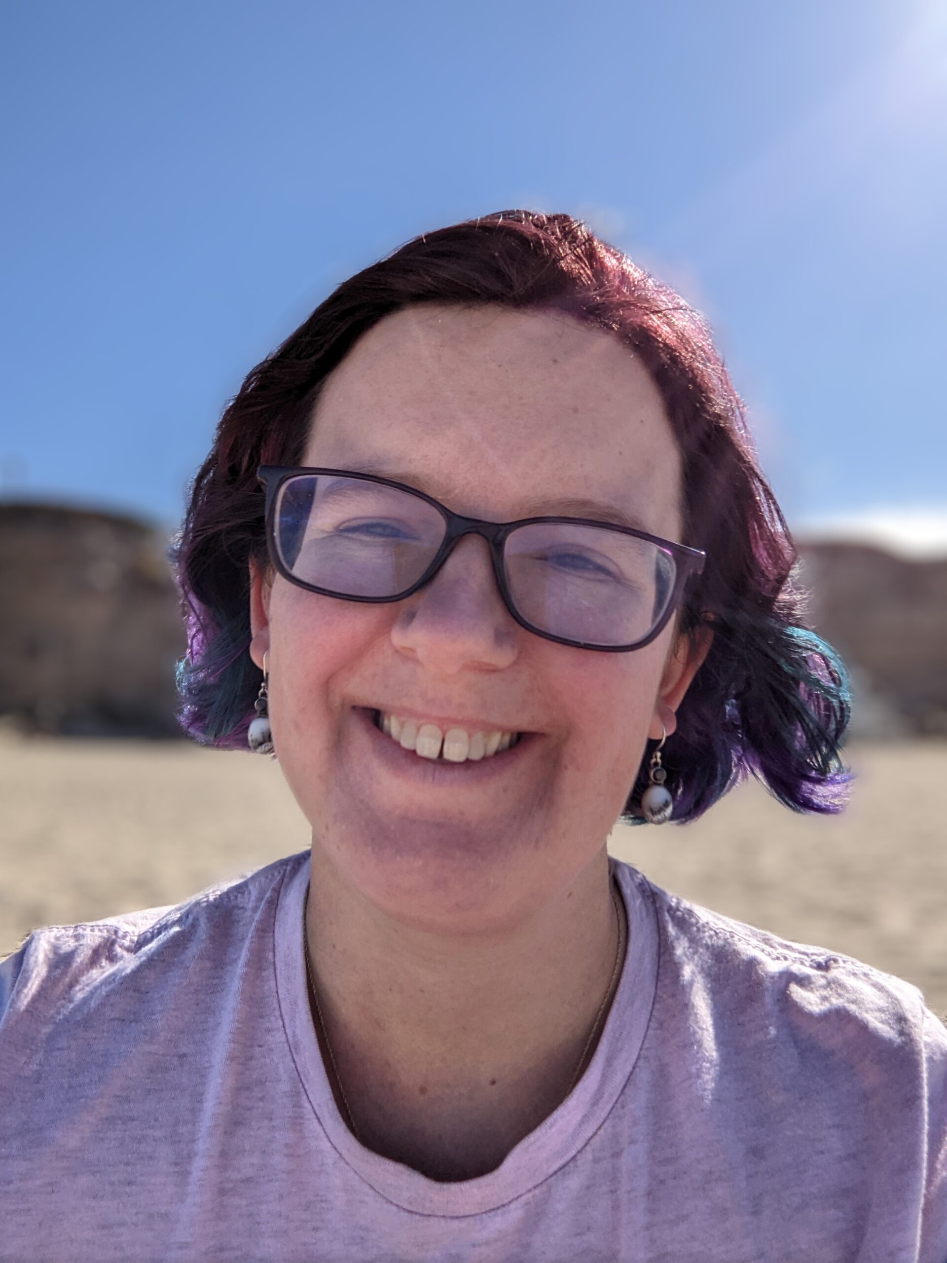 White woman with blue and brown hair. She's standing on the beach, wearing purple glasses and smiling widely.